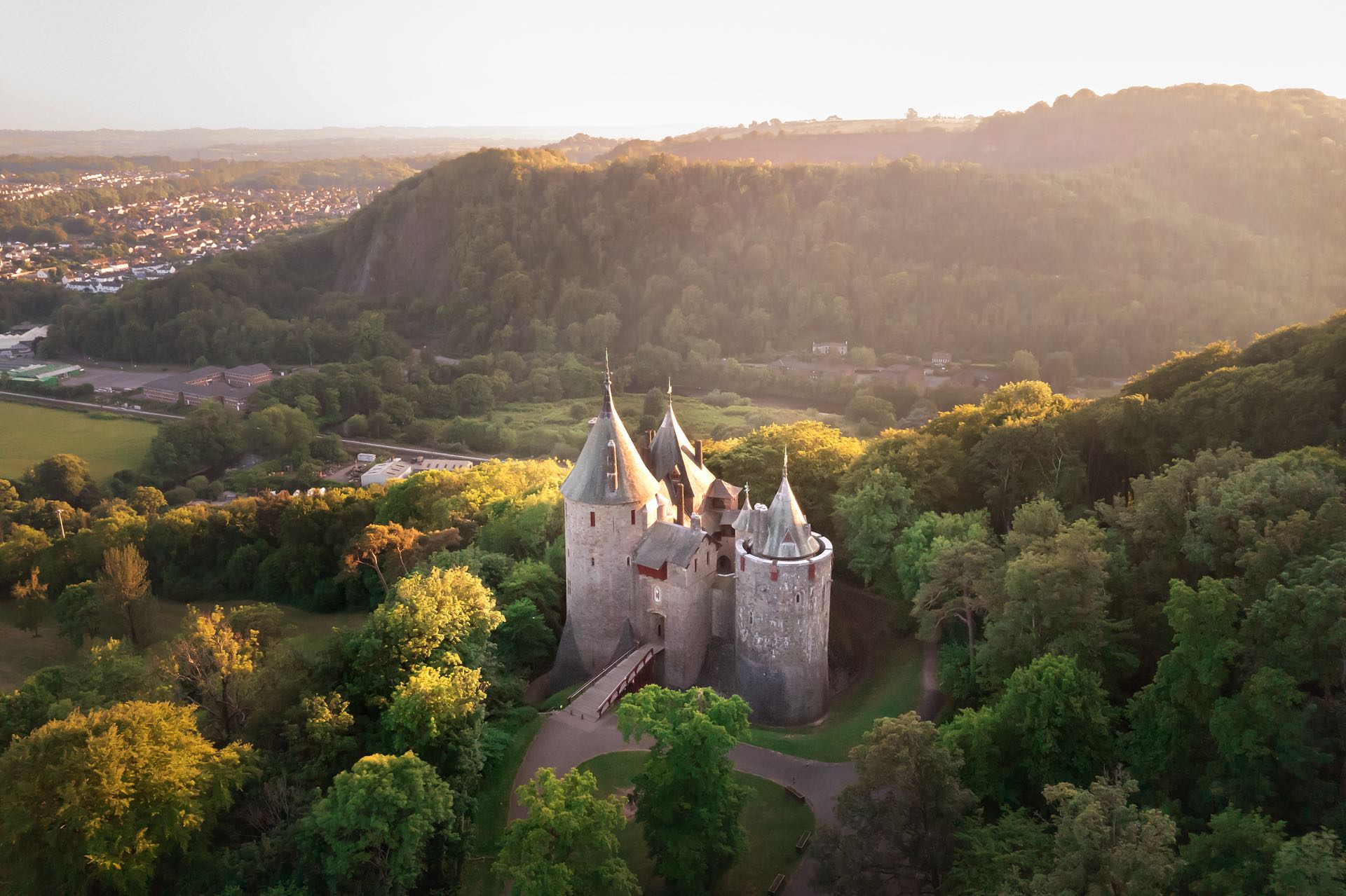 An aerial photograph of a medieval castle, with multiple conical towers, nestled in a dense, green, wooded valley during the golden hour, bathed in hazy golden sunlight. A bridge leads to the entrance, and the background shows continued forested hills and a small town. This image captures the castle from above.